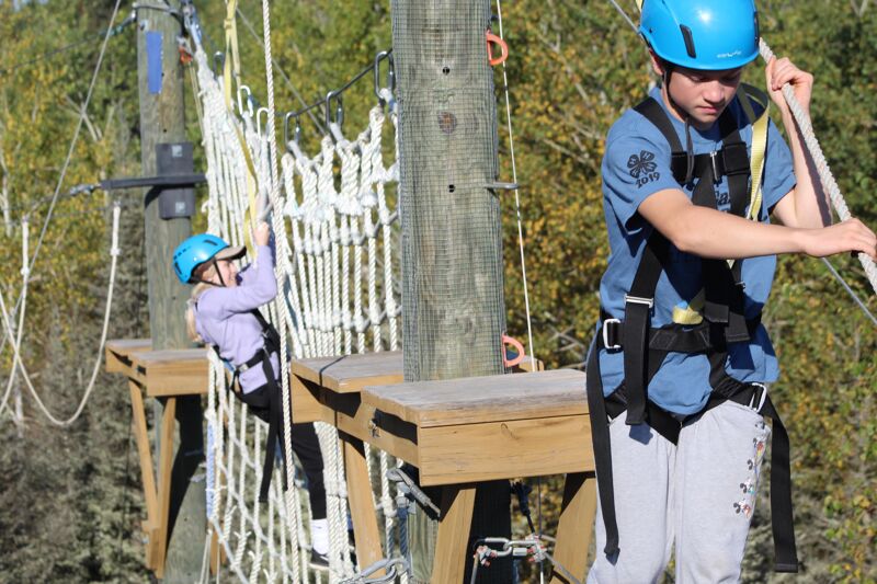 The image shows two people on a ropes course. A young girl is carefully navigating a rope bridge, secured with a harness and helmet. To the right, a young man, also wearing a helmet and harness, is holding onto a rope, possibly assisting or supervising. The course is set amidst trees, suggesting an outdoor adventure setting.